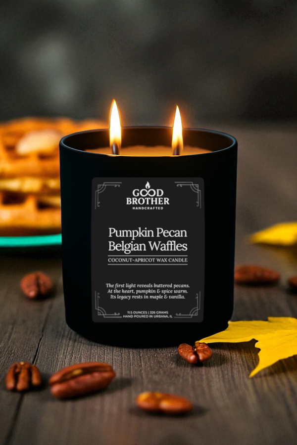 Black candle labeled 'Pumpkin Pecan Belgian Waffles'  by Good Brother with pumpkin pecan Belgian waffles on a wooden surface.