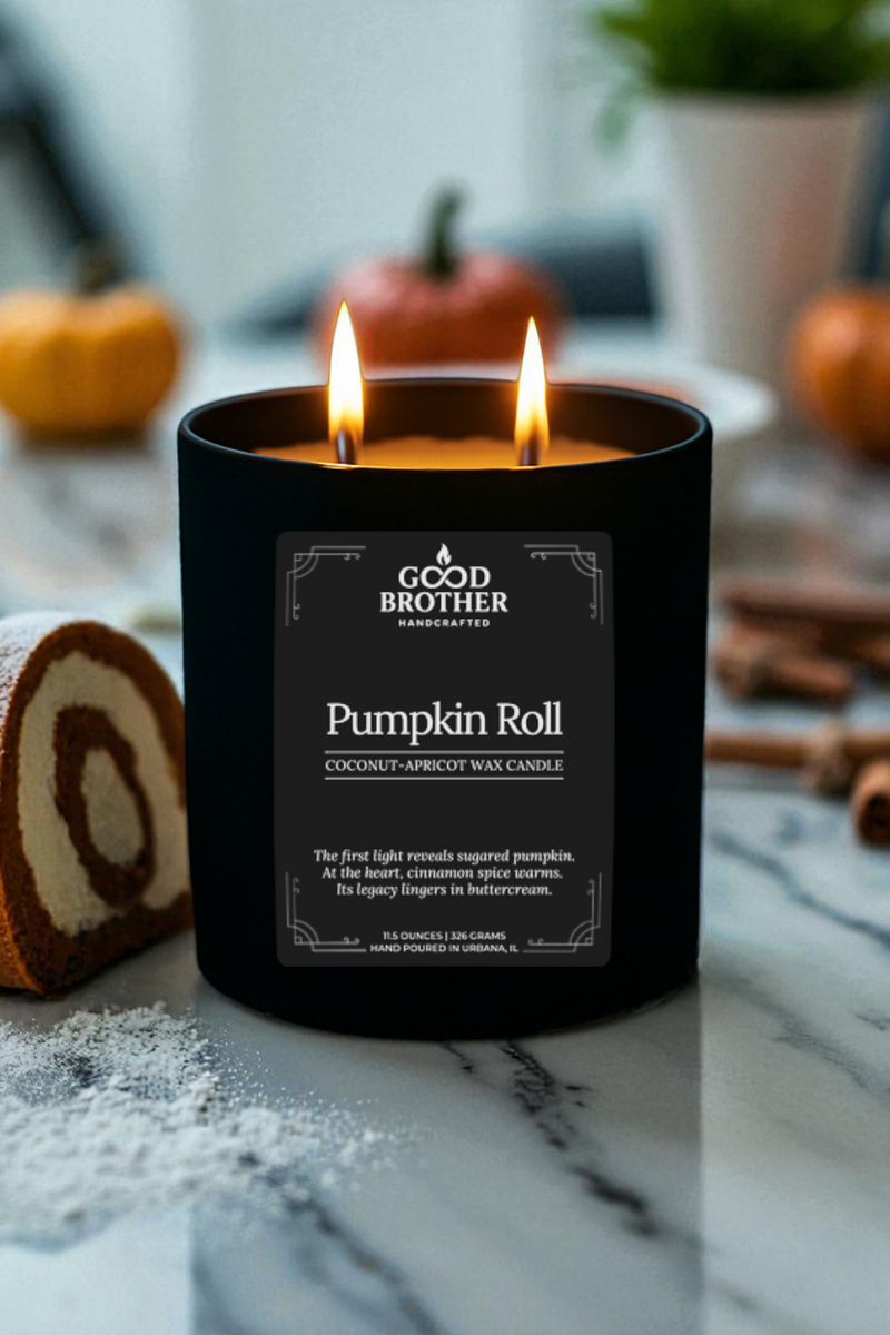Black candle labeled 'Pumpkin Roll' by Good Brother on a marble surface with  a slice of pumpkin roll cake and cinnamon sticks in the background.
