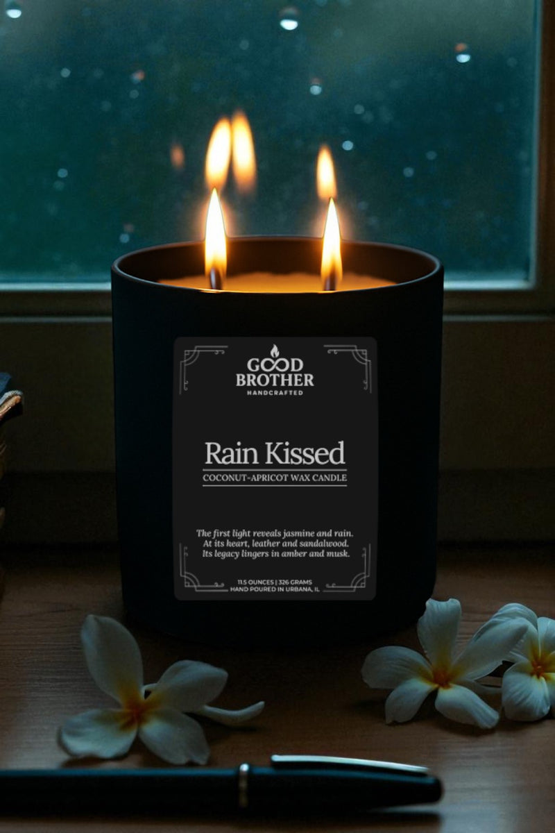 Candle labeled 'Rain Kissed' by Good Brother with flowers and a pen on a surface.