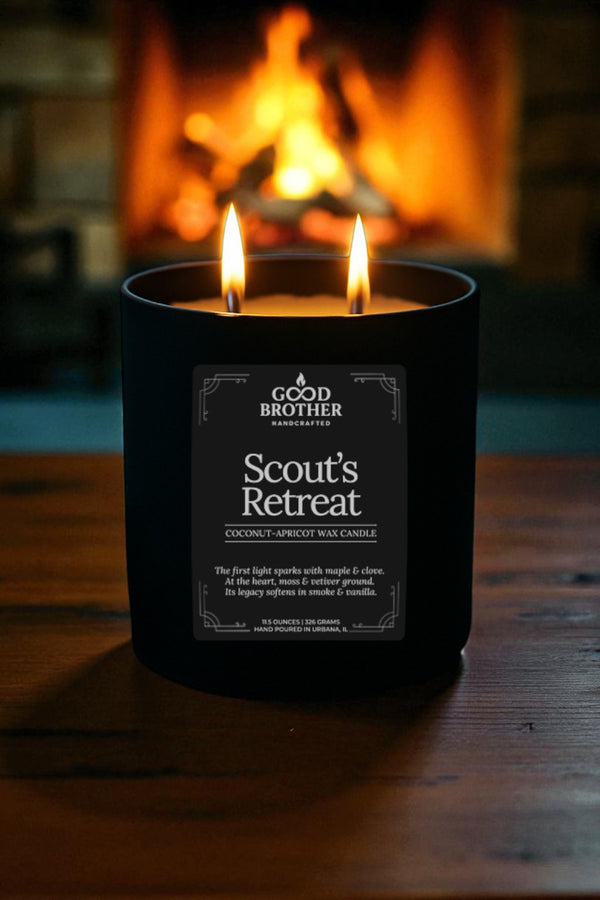 Black candle labeled 'Scout's Retreat' by Good Brother in front of a fireplace.