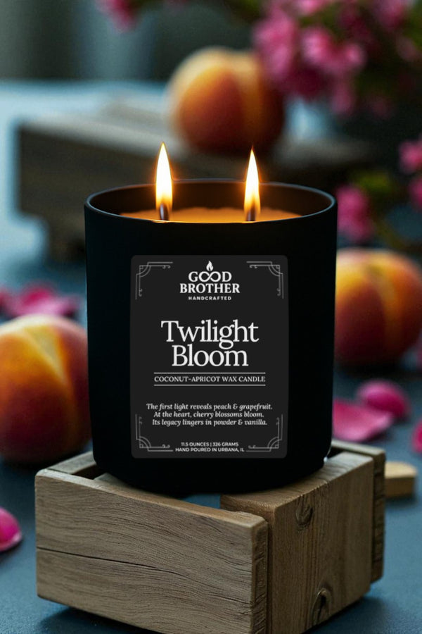 Black candle labeled 'Twilight Bloom' by Good Brother on a wooden stand with peaches and flowers in the background.